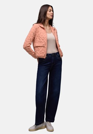 Femme portant une veste texturée rose saumon, un haut beige, un jean large bleu foncé et des baskets blanches, debout avec les mains derrière le dos.