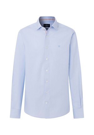 Hombre que lleva una camisa azul claro con botones, pantalones azul oscuro y zapatos de vestir negros, de pie con una mano en el bolsillo contra un fondo neutro.