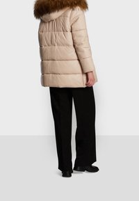 Puffjacka i ljusbeige med en huva i pälsimitation, med en kviltad design och långa ärmar, tillsammans med svarta vida byxor.