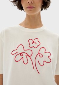 Vit bomullst-shirt med röd broderad blommönster med tre abstrakta blommor, rund halsringning och korta ärmar.