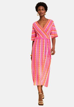 Naine kannab poolpikka wrap-kleiti roosa, oranži ja valge laineliste triipudega, mida täiendab rõngaskõrvarõngaste ja beežide kõrge kontsaga sandaalidega.