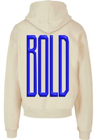 Felpa crema con cappuccio e maniche lunghe. Presenta una grande scritta "BOLD" blu sul retro in stile 3D. Morbida al tatto e dal taglio rilassato.