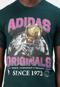 Green cotton t-shirt featuring a large graphic of a hand holding a golden trophy, "ADIDAS" and "ORIGINALS" printed in pink above.