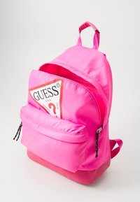 Rosa nylon ryggsekk med stor frontlomme, håndtak på toppen og glidelåser med svart og hvit stripestriper. Har et logo merke på fronten.