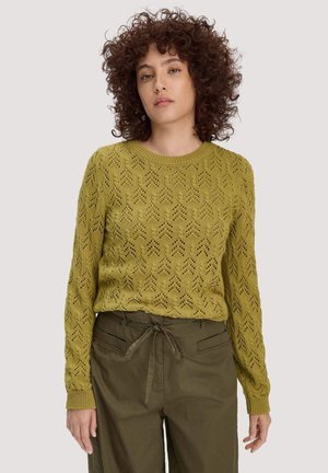 Frau mit lockigem Haar, die einen olivgrünen Pullover mit Muster und passende olivgrüne Jogginghosen mit Kordelzug trägt, steht vor einem schlichten Hintergrund.