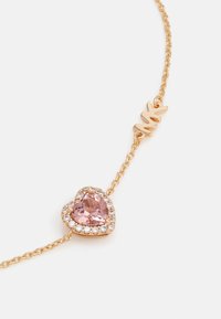 Pulsera de cadena de oro con una piedra rosa en forma de corazón rodeada de pequeñas gemas transparentes, y un cierre decorativo grabado con "MK."