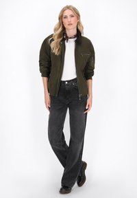 Olivgrüne Bomberjacke mit braunem Kragen, Reißverschlusstaschen, kombiniert mit hochgeschnittenen schwarzen Jeans und schwarzen Stiefeln. Glatte Textur.