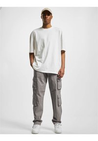 T-shirt blanc oversize, pantalon cargo gris clair avec multiples poches, et baskets blanches. Le mannequin porte une casquette de couleur claire.