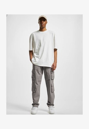 T-shirt blanc oversize, pantalon cargo gris clair avec multiples poches, et baskets blanches. Le mannequin porte une casquette de couleur claire.