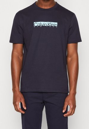 Muž nosící černé tričko s krátkým rukávem a světle modrým logem "Calvin Klein New York" přes hruď, doplněné černými kalhotami.