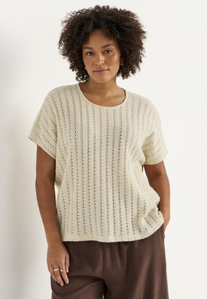 Kvinde med krøllet hår iført en løs cremefarvet kortærmet strikbluse og brune bukser, stående med den ene hånd i lommen mod en hvid baggrund.