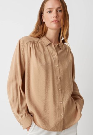 Chemise beige clair à boutons en tissu lisse, avec un col, des manches longues avec poignets, et de subtils détails de pli sur les épaules.