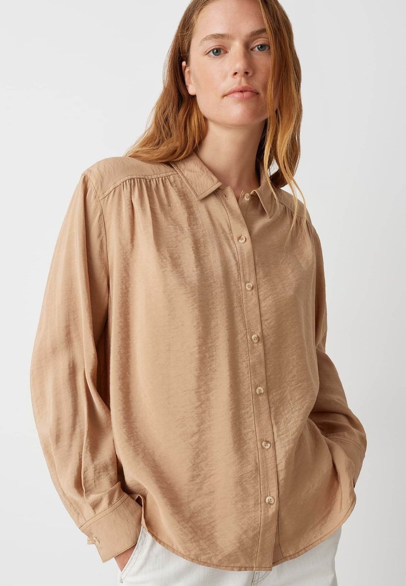 Camicia button-up beige chiaro in tessuto liscio, con colletto, maniche lunghe con polsini e delicati dettagli con pieghe sulle spalle.