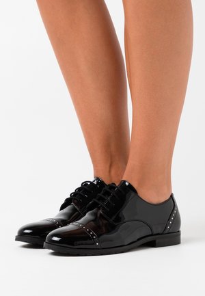 Lesklé černé lakované oxford boty s šněrovacím designem a dekorativními hřebíky nošené na holých nohách na bílém pozadí.