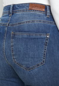 Jeans de ganga em lavagem azul escura com cintura alta, apresentando um rótulo da marca na parte de trás, costura em cobre e dois bolsos traseiros.