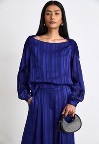 Lilla bluse med vertikale striper og puffermer, sammen med plisserte bukser. Modellen holder en svart, buet håndveske dekorert med edelstener.