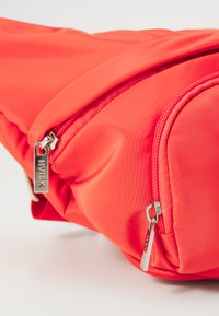 Crvena najlonska fanny paketa s glatkom teksturom, koja ima srebrne zatvarače na zip-u i detalje logotipa na povlačenju zatvarača. Kompaktan, trokutastog oblika.