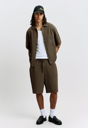 Young man with long hair wearing an olive green short-sleeve button-up shirt, matching shorts, white tank top, black cap, white socks, and black loafers.