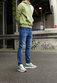 Zielona bluza z kapturem, niebieskie dżinsy i szare sneakersy Nike z turkusowymi akcentami. Gładkie faktury i swobodny krój, stoją na utwardzonej, miejskiej nawierzchni.