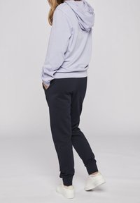 Svetle fialová mikina s kapucí a žebrovaným lemem, spárovaná s tmavě modrými tepláky s bočními kapsami. Boty jsou bílé s texturovanou podešví.