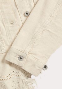 Giacca di denim crema con rifiniture ricamate, caratterizzata da una chiusura con bottoni a pressione argentati e dettagli di cucitura visibili sui polsini.
