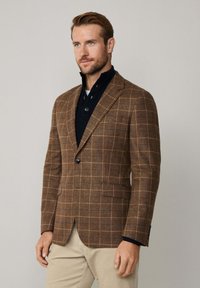 Braun karierter Blazer aus strukturiertem Material mit einer Knopfverschluss und Reverskragen, kombiniert mit einem schwarzen Stehkragen-Shirt darunter.