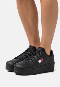 Czarne sneakersy na platformie z gładką skórzaną powierzchnią, zaokrąglonym noskiem i czarnymi sznurowadłami. Posiadają małą naszywkę z logo z czerwono-biało-niebieskimi akcentami.