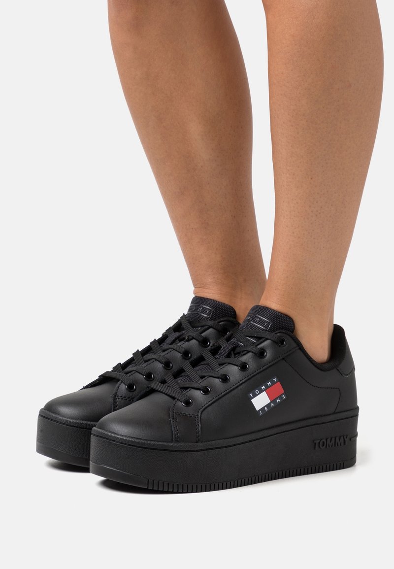 Czarne sneakersy na platformie z gładką skórzaną powierzchnią, zaokrąglonym noskiem i czarnymi sznurowadłami. Posiadają małą naszywkę z logo z czerwono-biało-niebieskimi akcentami.