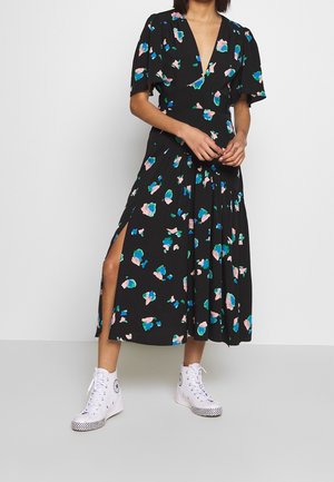 Vrouw draagt een zwarte midi-jurk met blauwe en roze bloemenprints, een schuine split aan de zijkant en witte hoge sneakers, staand tegen een effen achtergrond.