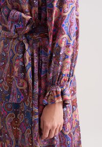 Bunt bedruckter Stoff im Paisley-Muster mit lila, pinken und blauen Farbtönen; hat einen gerafften Ärmel mit Rüschendetails und eine gebundene Taille.