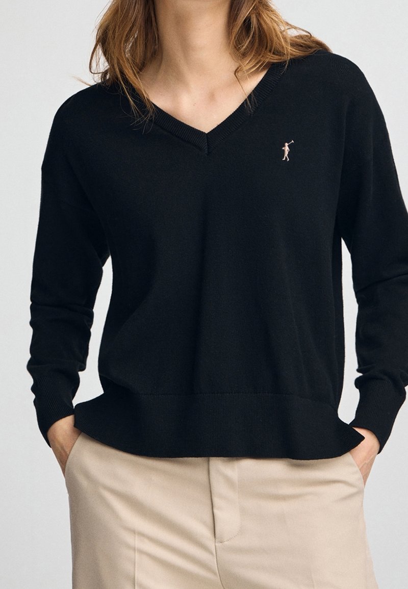 Femme portant un pull noir à col en V avec une petite figure brodée sur la poitrine et un pantalon beige, les mains dans les poches.