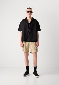 Camicetta nera a maniche corte con texture sopra una canotta nera, abbinata a pantaloni cargo beige chiaro e sneakers nere con suola bianca.