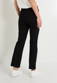 Persona vistiendo pantalones negros ajustados y zapatillas deportivas blancas, de pie contra un fondo blanco liso, con una camiseta blanca parcialmente visible.