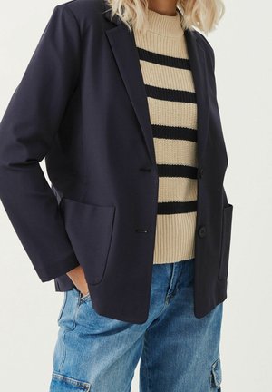 Person trägt ein schwarzes Blazer über einem beige-schwarz gestreiften Pullover mit blauer Jeans-Cargohose, Hand in der Tasche, steht vor einem weißen Hintergrund.