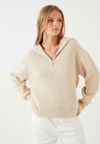 Beige stickad tröja med dragkedja i halsen och vida ärmar. Har en texturerad stickad mönster och ribbade mudd och fåll.