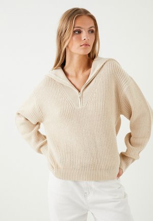 Suéter de punto beige con cuello con cremallera y mangas anchas. Presenta un patrón de puntada texturizada y puños y dobladillo acanalados.