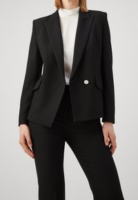 Černý blazer s hladkými klopami, zapínání na jeden stříbrný knoflík, dvě přední kapsy a hladká tkanina, spárovaný s bílým topem s vysokým límcem.