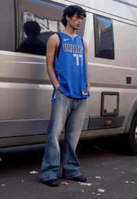 Maillot de basketball bleu avec "DALLAS" et le numéro "77," associé à un jean en denim bleu clair ample et des chaussures noires à enfiler.