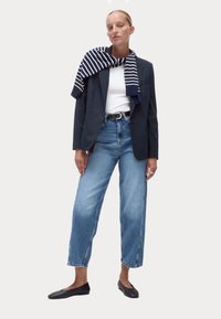 Blazer navy sur un t-shirt blanc, associé à un jean large bleu clair et des ballet flats noires. Une écharpe rayée bleu marine et blanche drape le cou.
