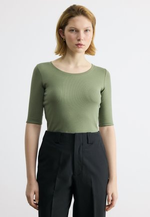 Młoda kobieta o krótkich blond włosach, ubrana w dopasowany zielony top z półdługimi rękawami i czarne spodnie z wysokim stanem, stojąca na białym tle.