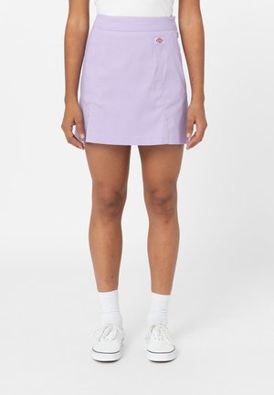 Person iført et lyse lilla A-formet skørt, hvide sokker og hvide snøresneakers, der står mod en ensfarvet baggrund.