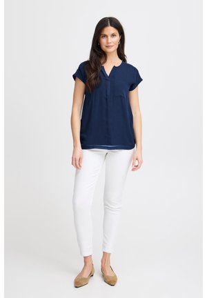 Woman standing, wearing a navy blue short-sleeve blouse, white slim pants, and tan flats, against a plain light background.