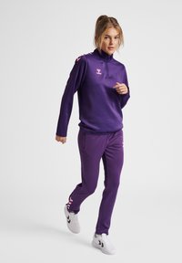 Survêtement de sport violet avec un haut zippé à col montant et des accents roses, associé à un pantalon fuselé assorti et des baskets blanches.