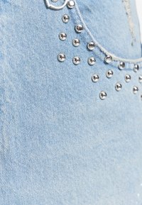 Tissu en denim bleu clair avec des clous ronds en métal argenté disposés près d'un bord effiloché et un détail de bague en métal.