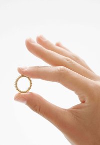 Anillo de oro con un diseño texturizado y con picos, sostenido entre dedos contra un fondo claro. La forma es circular con un centro abierto.