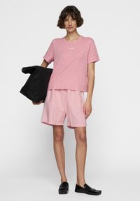 Pink kortærmet T-shirt med tekstureret stof, parret med lyserøde shorts og sorte slip-on sko, mens der holdes en sorte taske.