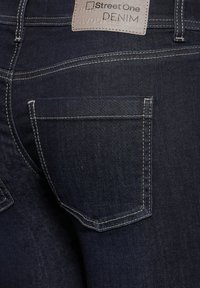 Un jean en denim bleu foncé présente une poche arrière bien visible avec des coutures blanches et une étiquette en cuir beige affichant "Street One Denim."