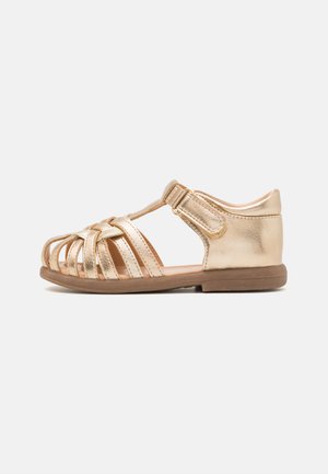 Gullmetallisk sandal med åpen tå-design, flere flettede stropper, justerbar ankelreim og en flat, brun såle.