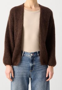 Brauner, flauschiger Cardigan mit offenem Vorderteil und langen, ballonartigen Ärmeln über einem beigen Top und blauen hoch taillierten Jeans. Weiche Textur und lockere Passform.