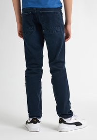 Jeans de mezclilla de color azul oscuro con un corte slim, que presentan bolsillos traseros con detalles de costura en forma de triángulo. Combinados con zapatillas blancas.